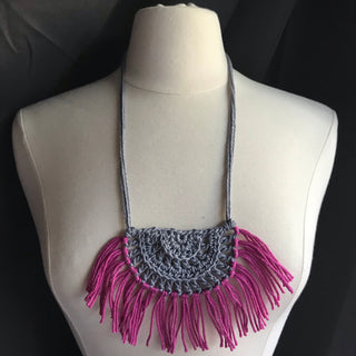 Necklace made of yarn being worn by model form figure.  Necklace color is a light gray called Silver for the tie and half circle with the fringe being a jewel tone Pink called Rose.  Necklace design is half circle made with textured stitches and fringe hanging from the edge around.