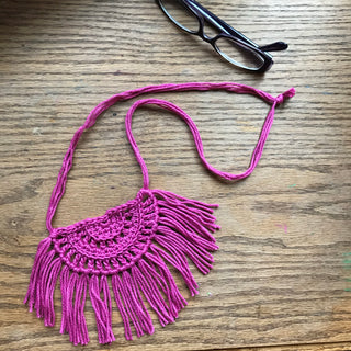 Necklace made of yarn laying on a wooden flat surface next to a pair of glasses.  Necklace color is a jewel tone Pink called Rose.  Necklace design is half circle made with textured stitches and fringe hanging from the edge around.