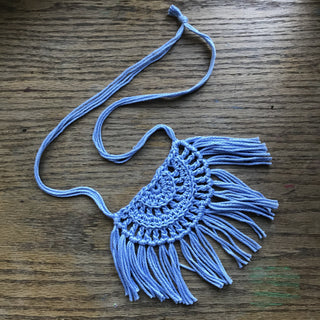Necklace made of yarn laying on a wooden flat surface.  Necklace color is a light blue called Sky.  Necklace design is half circle made with textured stitches and fringe hanging from the edge around.