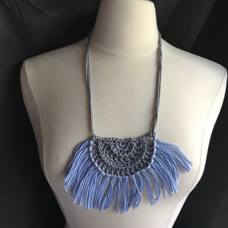 Necklace made of yarn being worn by model form figure.  Necklace color is a light gray called Silver for the tie and half circle with the fringe being a light blue called Sky.  Necklace design is half circle made with textured stitches and fringe hanging from the edge around.