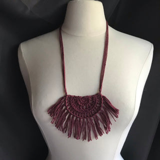 Necklace made of yarn being worn by model form figure.  Necklace color is a reddish/burgundy called Merlot.  Necklace design is half circle made with textured stitches and fringe hanging from the edge around.