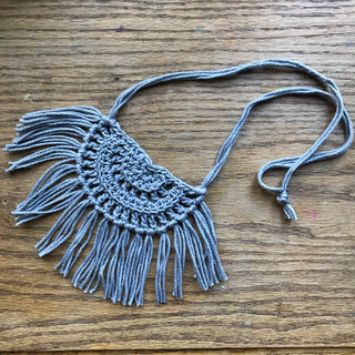 Necklace made of yarn laying on a wooden flat surface.  Necklace color is a light gray called Silver.  Necklace design is half circle made with textured stitches and fringe hanging from the edge around.