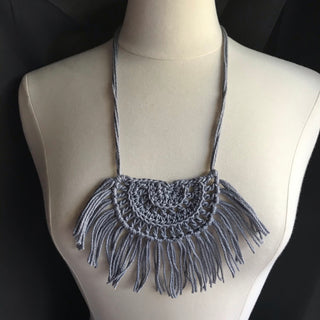 Necklace made of yarn being worn by model form figure.  Necklace color is a light gray called Silver.  Necklace design is half circle made with textured stitches and fringe hanging from the edge around.