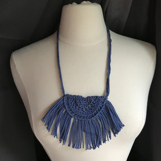 Necklace made of yarn being worn by model form figure.  Necklace color is a jewel tone Blue called Denim.  Necklace design is half circle made with textured stitches and fringe hanging from the edge around.
