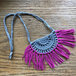 Necklace made of yarn laying on a wooden flat surface.  Necklace color is a light gray called Silver for the tie and half circle with the fringe being a jewel tone Pink called Rose.  Necklace design is half circle made with textured stitches and fringe hanging from the edge around.