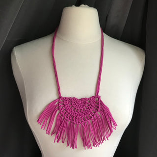 Necklace made of yarn being worn by model form figure.  Necklace color is a jewel tone Pink called Rose.  Necklace design is half circle made with textured stitches and fringe hanging from the edge around.