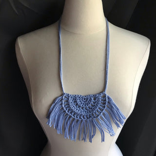Necklace made of yarn being worn by model form figure.  Necklace color is a light blue called Sky.  Necklace design is half circle made with textured stitches and fringe hanging from the edge around.
