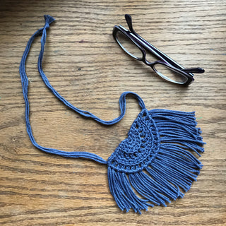 Necklace made of yarn laying on a wooden flat surface next to a pair of glasses.  Necklace color is a jewel tone Blue called Denim.  Necklace design is half circle made with textured stitches and fringe hanging from the edge around.