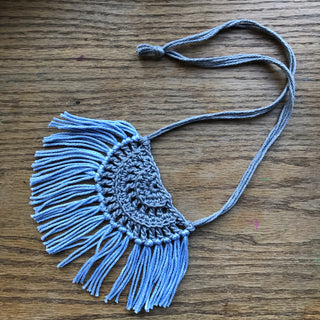 Necklace made of yarn laying on a wooden flat surface.  Necklace color is a light gray called Silver for the tie and half circle with the fringe being a light blue called Sky.  Necklace design is half circle made with textured stitches and fringe hanging from the edge around.